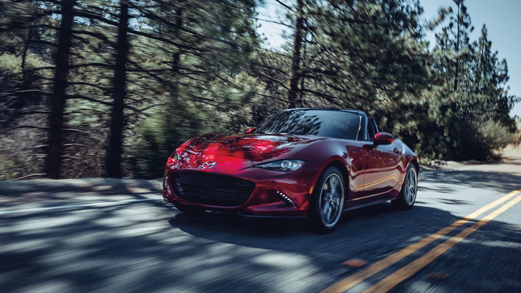 Red 2023 Mazda MX-5 Miata drives down forest road.